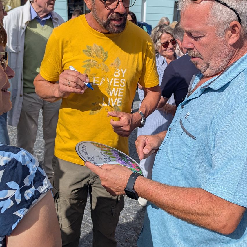 Ein Mann in einem gelben T-Shirt unterschreibt ein Poster mit einem Stift. Ein älterer Mann in einem blauen Hemd steht vor ihm. Sie sind draußen.