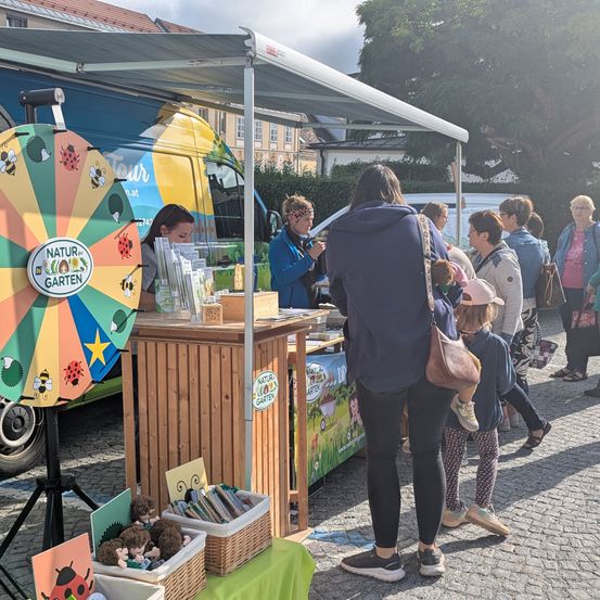 Eine Gruppe von Menschen steht an einem Marktstand mit Büchern und Spielzeug im Angebot. Eine Frau mit Handtasche steht vor einem Tisch mit einem bunten Rad. Leute gehen in der Nähe vorbei.