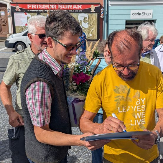 Ein Mann mit Brille unterschreibt ein Blatt Papier vor einer Gruppe von Menschen draußen. Hinter ihm schaut ein Mann in einem karierten Hemd und Weste zu.