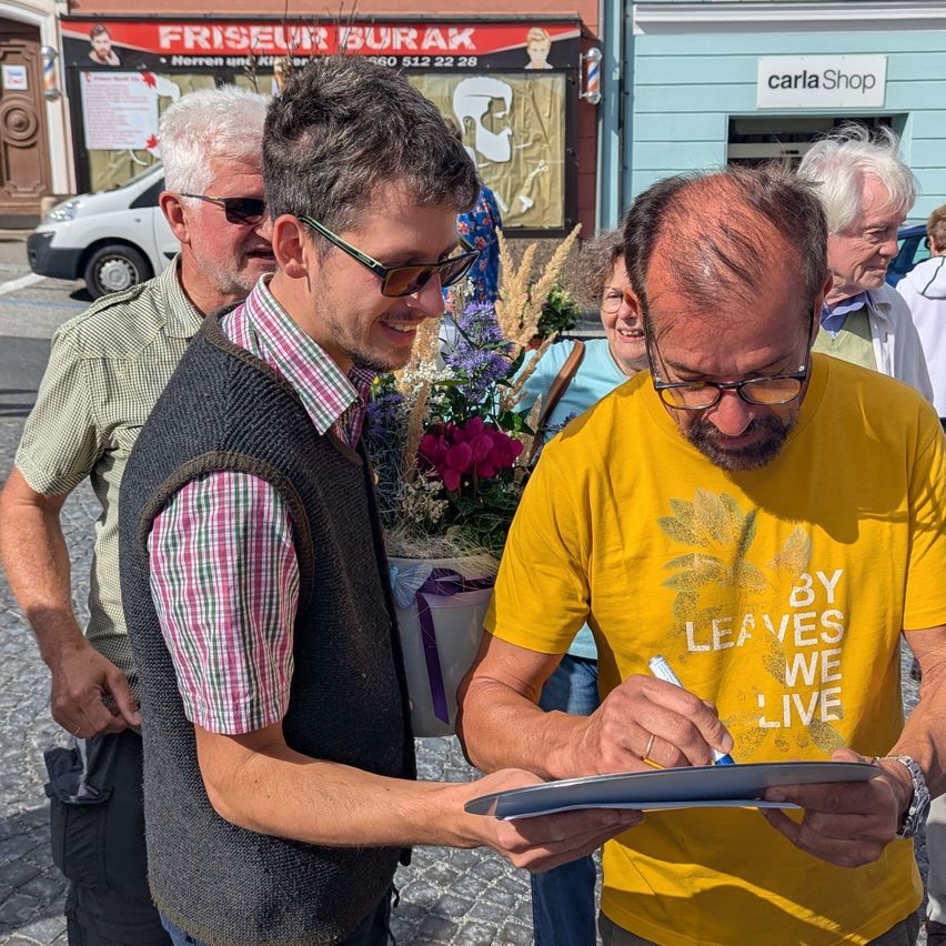 Ein Mann mit Brille unterschreibt ein Blatt Papier vor einer Gruppe von Menschen draußen. Hinter ihm schaut ein Mann in einem karierten Hemd und Weste zu.