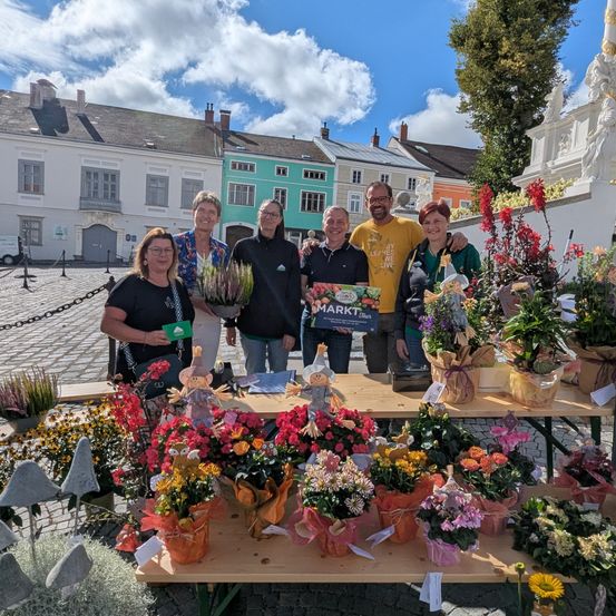 Mehrere Personen stehen hinter einem Tisch mit Blumenarrangements auf einem Platz. Hinter ihnen befinden sich mehrere Gebäude. Ein Mann hält ein Schild mit der Aufschrift 'MARKT'.