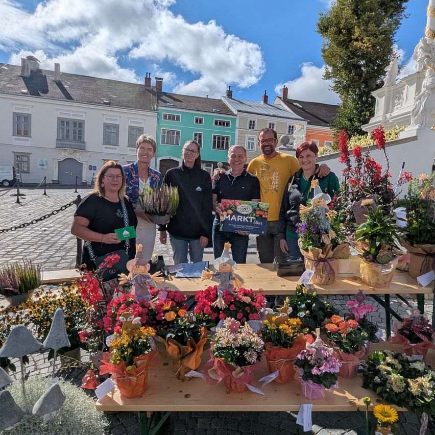 Mehrere Personen stehen hinter einem Tisch mit Blumenarrangements auf einem Platz. Hinter ihnen befinden sich mehrere Gebäude. Ein Mann hält ein Schild mit der Aufschrift 'MARKT'.