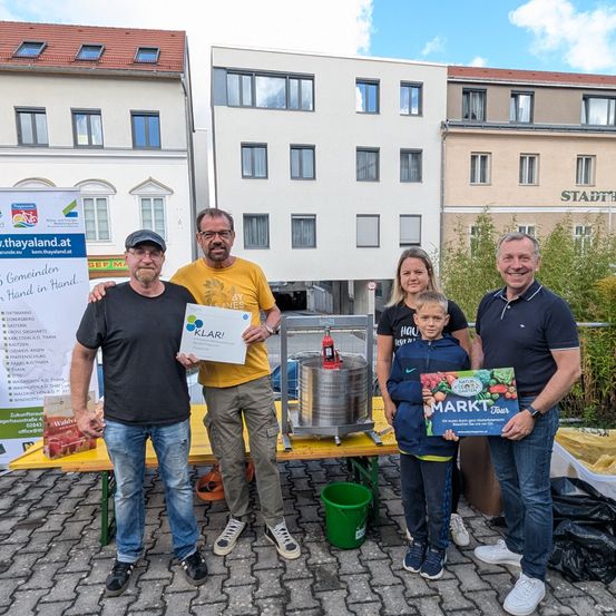 Eine Gruppe von Erwachsenen und einem Kind posiert lächelnd vor einem Tisch. Sie halten eine Box und ein Zertifikat. Hinter ihnen stehen Gebäude und ein Feuerlöscher.