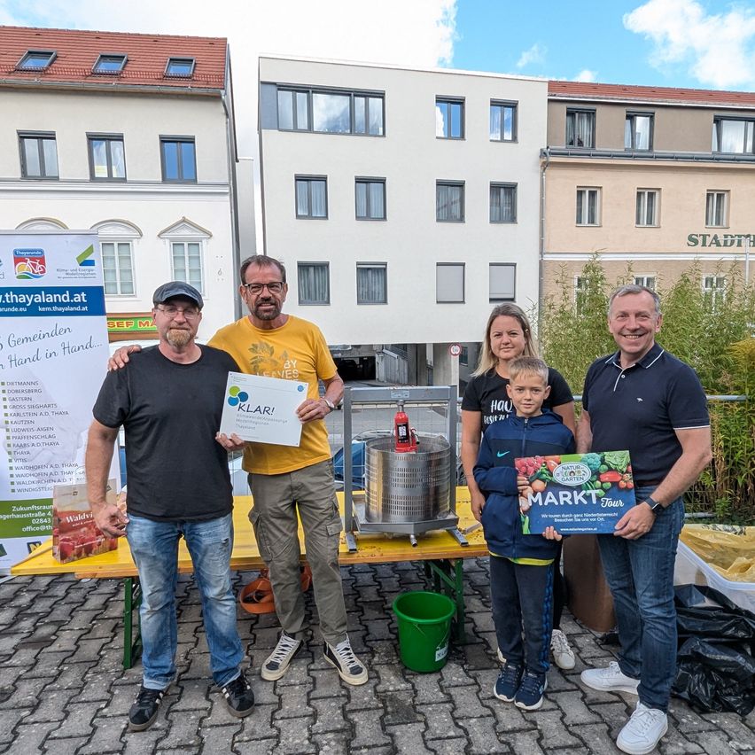 Eine Gruppe von Erwachsenen und einem Kind posiert lächelnd vor einem Tisch. Sie halten eine Box und ein Zertifikat. Hinter ihnen stehen Gebäude und ein Feuerlöscher.