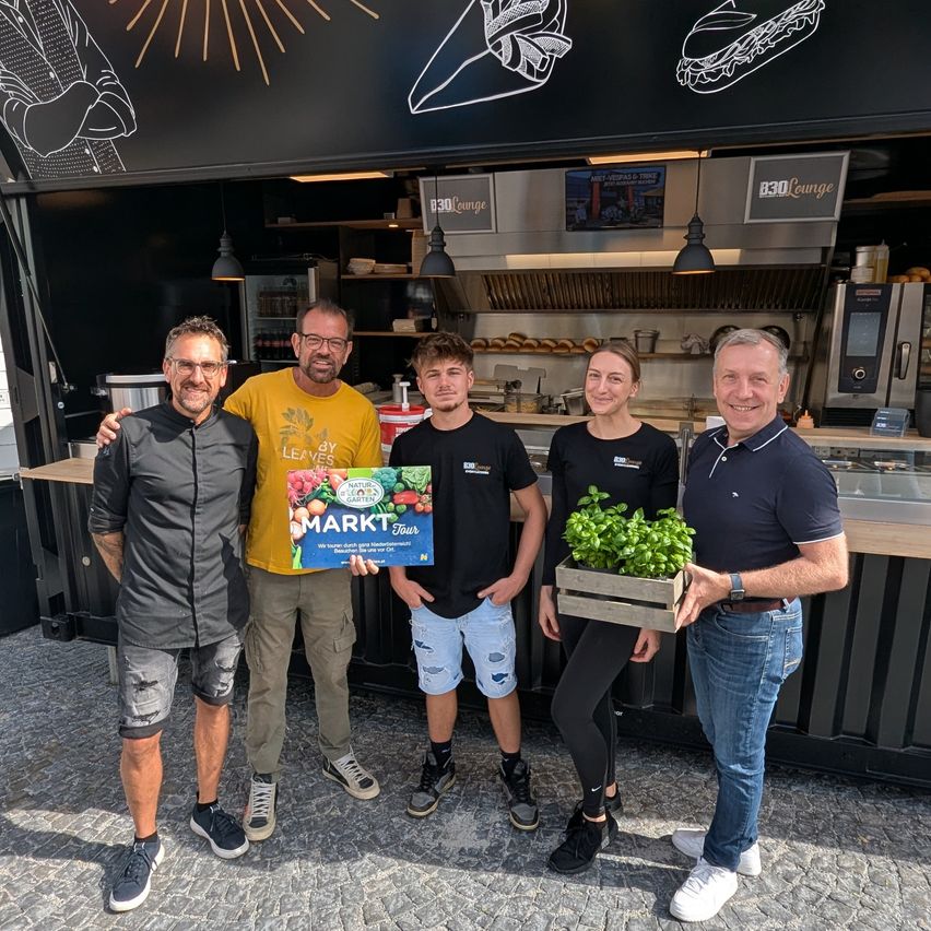 Fünf Personen stehen vor einem Imbisswagen, lächeln und posieren für ein Foto. Ein Mann hält eine blaue Box und eine Frau eine Topfpflanze. Dahinter ist eine Theke mit Essensartikeln zu sehen.