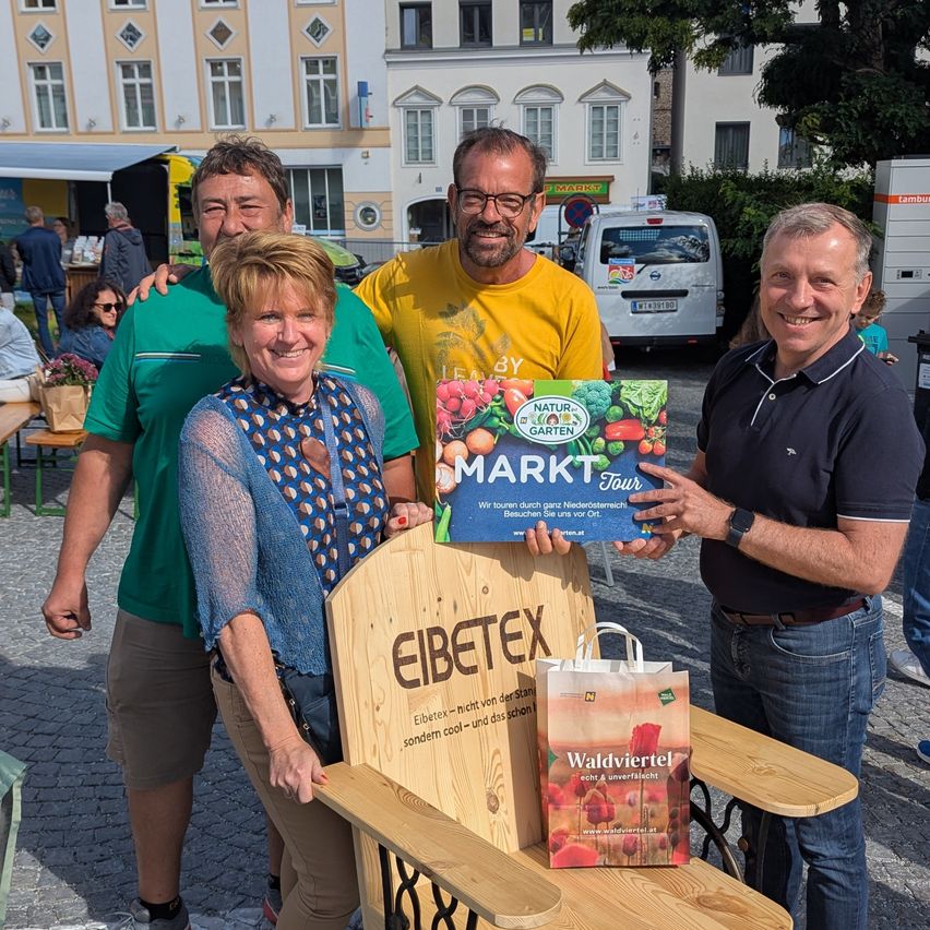 Vier Personen posieren für ein Foto bei einer Outdoor-Veranstaltung. Der Mann links trägt ein grünes Shirt, während die Frau in der Mitte eine gemusterte Bluse trägt. Der Mann rechts hält ein Banner und eine Tasche. Ein weißer Van ist hinter ihnen geparkt.
