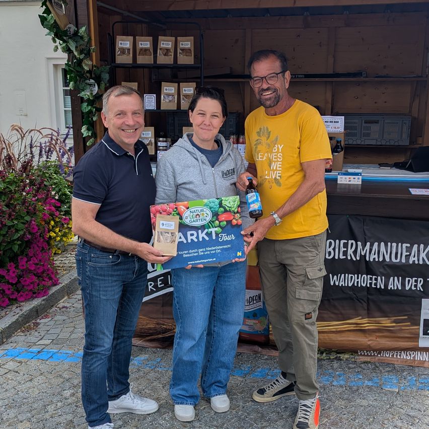 Drei Menschen stehen lächelnd vor einem Stand. Ein Mann hält eine Flasche und ein Buch, ein anderer Mann hält eine Schachtel und eine Frau hält eine Schachtel. Dahinter befindet sich ein Banner.