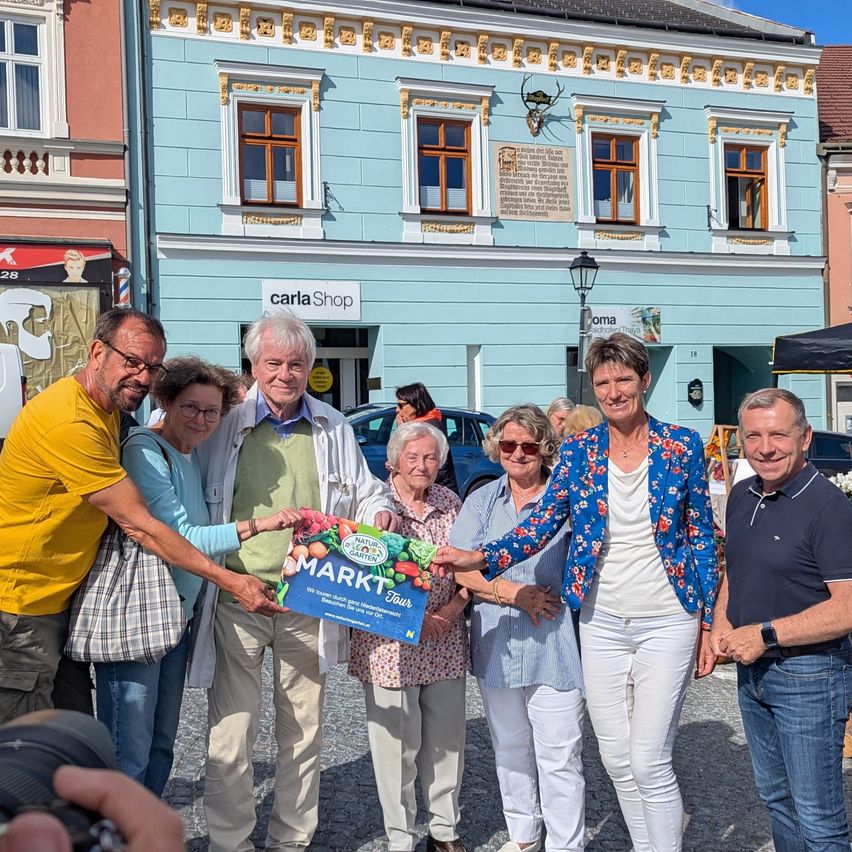 Eine Gruppe älterer Menschen steht vor einem blauen Gebäude und hält eine Box mit der Aufschrift MARKT Tour. Sie lächeln und posieren für ein Foto. Ein Mann mit einer Kamera macht ihr Bild. Dahinter steht ein Gebäude mit einer Lampe und einem Schild, das Carla Shop besagt.