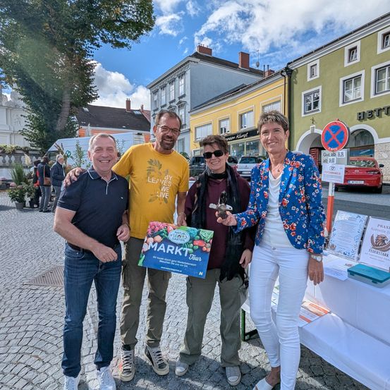 Vier Personen stehen auf einer Kopfsteinpflasterstraße und lächeln. Ein Mann hält ein Schild mit der Aufschrift 'Markt Tour'. Eine Frau hält einen kleinen Gegenstand. Hinter ihnen steht ein Tisch mit Broschüren. Im Hintergrund befinden sich Gebäude und Bäume.