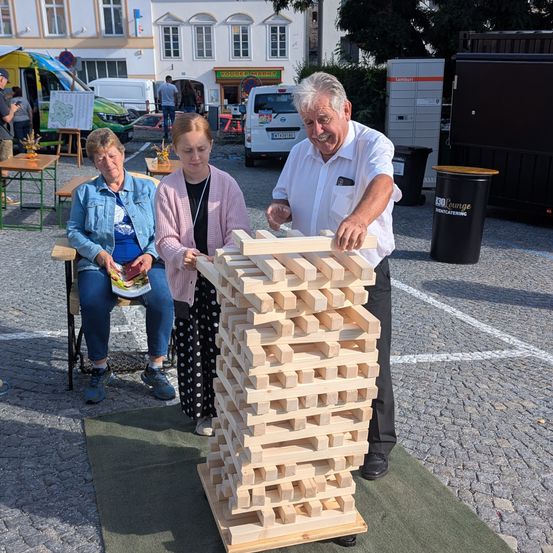 Ein älterer Mann baut einen Turm aus Holzklötzen, während zwei Frauen zusehen. Sie sind draußen in der Nähe eines Gebäudes und einiger geparkter Fahrzeuge.