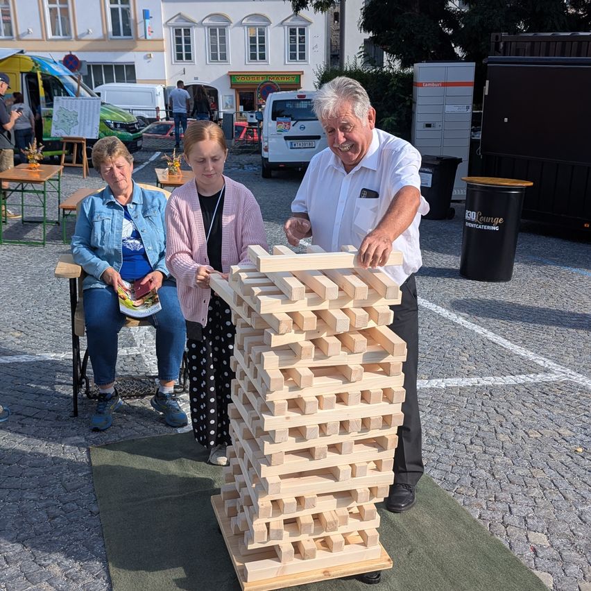 Ein älterer Mann baut einen Turm aus Holzklötzen, während zwei Frauen zusehen. Sie sind draußen in der Nähe eines Gebäudes und einiger geparkter Fahrzeuge.
