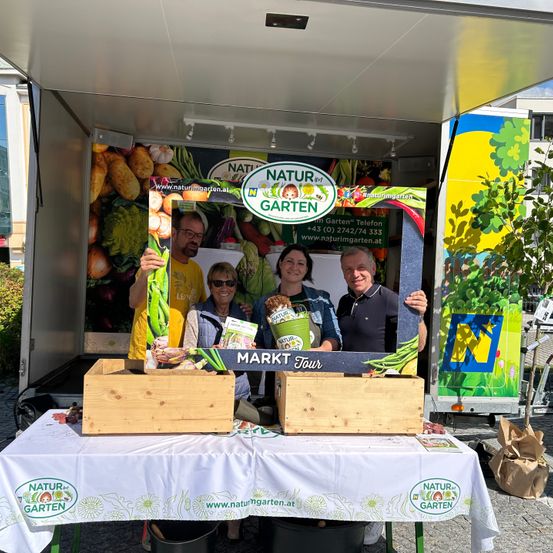 Vier Personen stehen vor einem Lebensmittelwagen mit dem Schild 'Markt Tour' und Holzkisten voller Gemüse. Ein Schild mit 'Natur im Garten' ist hinter ihnen.