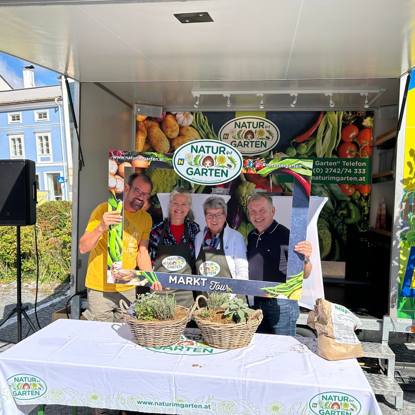 Vier Personen stehen in einem mobilen Laden und halten ein Schild mit der Aufschrift 'Markt Tour'. Sie lächeln und scheinen für ein Foto zu posieren. Vor ihnen stehen Körbe mit Pflanzen auf dem Tisch.