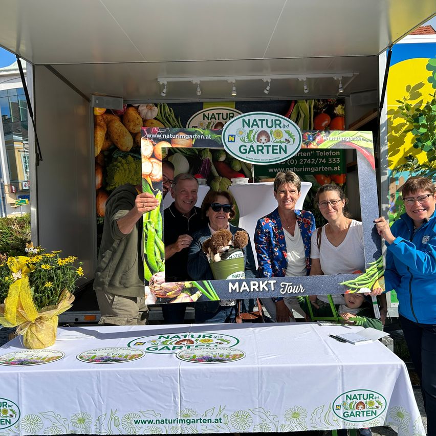 Fünf Personen posieren für ein Foto vor einer Anzeigetafel mit dem Schriftzug 'NATUR MARKT Tour'. Die Tafel zeigt Gemüse und einen Plüschtier. Sie stehen neben einem Tisch mit weißer Tischdecke und einem Blumenstrauß. Dahinter steht ein Fahrzeug mit einem großen Bild von Gemüse auf der Seite.