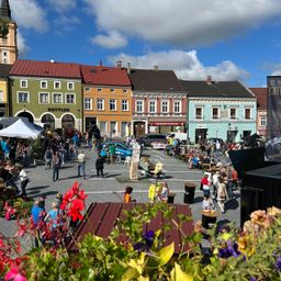 Ein geschäftiger Stadtplatz mit vielfältigen, farbenfrohen Gebäuden. Viele Menschen versammeln sich, essen und erkunden das Gebiet, mit einem Zelt, einem Banner und geparkten Autos.