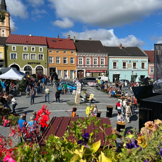 Ein geschäftiger Stadtplatz mit vielfältigen, farbenfrohen Gebäuden. Viele Menschen versammeln sich, essen und erkunden das Gebiet, mit einem Zelt, einem Banner und geparkten Autos.