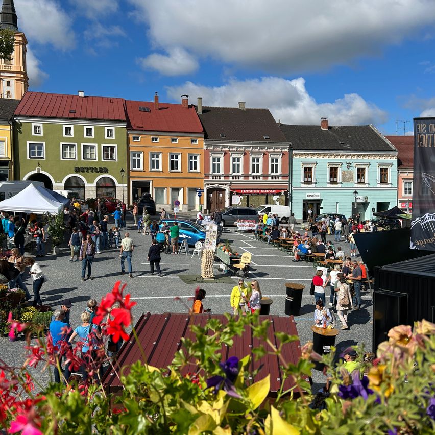 Ein geschäftiger Stadtplatz mit vielfältigen, farbenfrohen Gebäuden. Viele Menschen versammeln sich, essen und erkunden das Gebiet, mit einem Zelt, einem Banner und geparkten Autos.