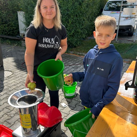 Eine Frau und ein Junge stehen gemeinsam neben einer Apfelpresse und halten grüne Eimer. Die Frau trägt ein schwarzes T-Shirt mit einem Hauslogo. Der Junge trägt eine blaue Kapuzenjacke. Sie sind umgeben von einem Tisch, einem weißen Auto und Grünpflanzen.