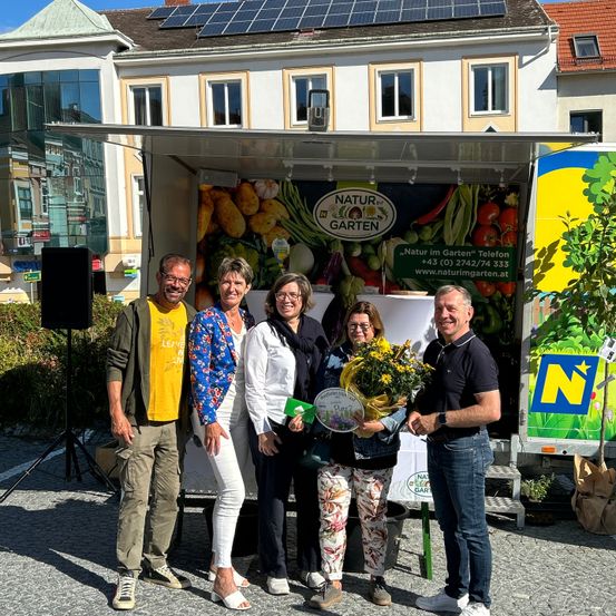 Fünf Personen stehen vor einem Lastwagen mit dem Schriftzug Natur im Garten. Eine Frau hält einen Blumenstrauß und eine grüne Karte. Hinter ihnen ist ein Gebäude mit Solarpanelen auf dem Dach zu sehen.