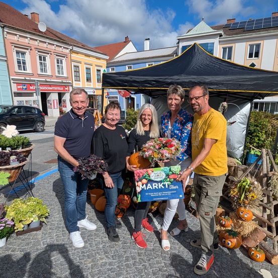 Fünf Personen stehen lächelnd unter einem Zelt, Blumen und eine Box haltend. Sie sind von Pflanzen, Kürbissen und Körben umgeben. Hinter ihnen sind Gebäude und Autos zu sehen.