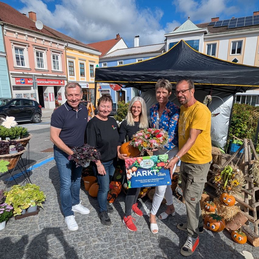 Fünf Personen stehen lächelnd unter einem Zelt, Blumen und eine Box haltend. Sie sind von Pflanzen, Kürbissen und Körben umgeben. Hinter ihnen sind Gebäude und Autos zu sehen.