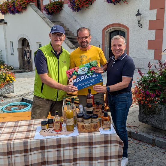 Drei Männer stehen lächelnd neben einem Tisch. Einer hält ein Buch, ein anderer zeigt eine Flasche und der dritte trägt eine Uhr. Sie sind draußen, in der Nähe eines Gebäudes mit Treppen und Blumen.
