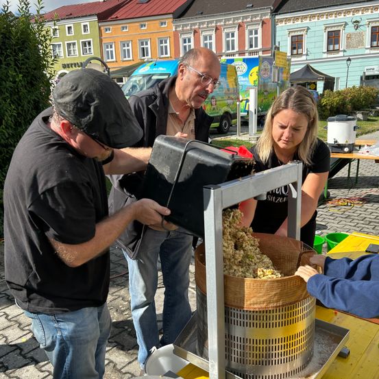 Drei Personen benutzen eine mechanische Traubenpresse im Freien. Gebäude und ein Van sind im Hintergrund zu sehen.
