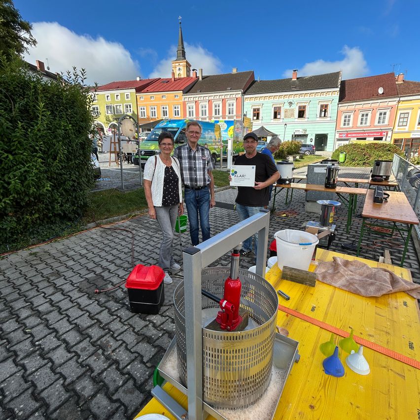 Drei Personen stehen neben einem gelben Tisch auf einem Stadtplatz, eine hält ein Schild. Im Hintergrund sind farbenfrohe Gebäude zu sehen.