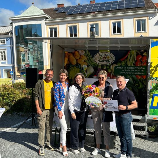 Fünf Personen stehen vor einem Truck, zwei Frauen halten einen Blumenstrauß und eine Frau hält ein Zertifikat. Sie lächeln und posieren für ein Foto. Hinter ihnen zeigt ein Schild den Text 'Natur Garten'. Ein Lautsprecher und eine grüne Pflanze sind auf der linken Seite und ein gelbes Schild ist auf der rechten Seite.