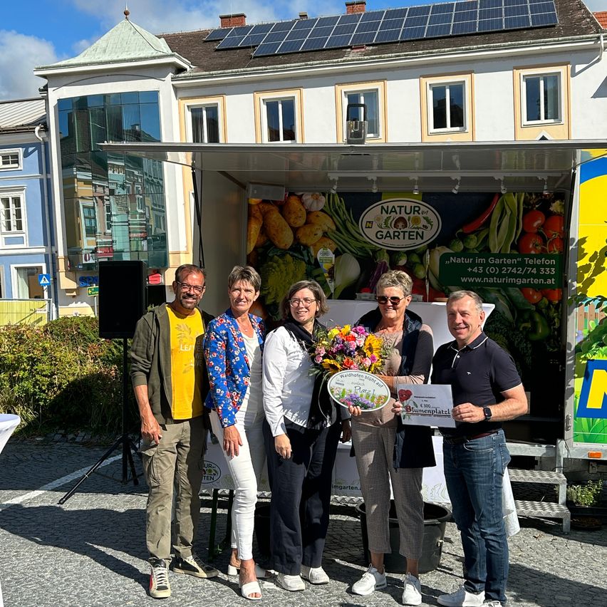 Fünf Personen stehen vor einem Truck, zwei Frauen halten einen Blumenstrauß und eine Frau hält ein Zertifikat. Sie lächeln und posieren für ein Foto. Hinter ihnen zeigt ein Schild den Text 'Natur Garten'. Ein Lautsprecher und eine grüne Pflanze sind auf der linken Seite und ein gelbes Schild ist auf der rechten Seite.