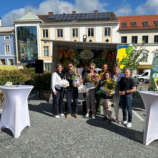 Eine Gruppe von Menschen mit Blumen und einem Schild steht vor einem Lastwagen mit bunter Dekoration auf einem Stadtplatz. Hinter ihnen sind Gebäude mit Solarmodulen zu sehen.