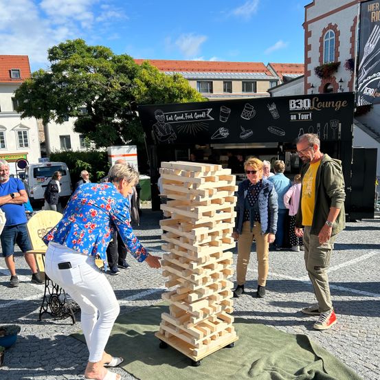 Eine Gruppe von Menschen spielt ein Jenga-Spiel auf einer Straße. Eine Frau in einer Bluse mit Blumenmuster lehnt sich vor, um einen Block zu entfernen. Hinter ihr beobachten zwei Männer, einer mit Brille und Uhr. Ein Eiswagen ist in der Nähe geparkt.