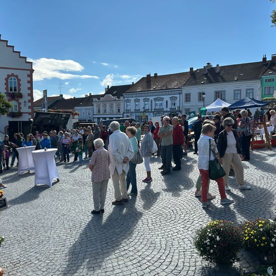 Viele Menschen versammeln sich auf einem Stadtplatz, einige mit Hüten und Brillen, vor Gebäuden mit Schornsteinen und Blumen. Es gibt weiße Tische und Zelte.