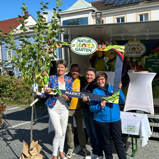 Vier Personen stehen vor einem Stand, der mit Natur im Garten beschriftet ist. Sie halten ein Schild mit der Aufschrift Markt Tour und posieren neben einem jungen Baum. Dahinter ist ein Tisch mit einer weißen Tischdecke und einem Banner zu sehen.