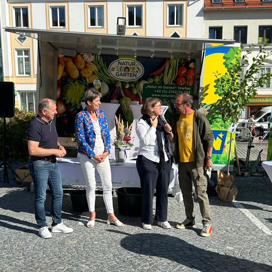 Vier Personen stehen vor einem Stand, der 'Natur im Garten' bewirbt. Eine Frau spricht in ein Mikrofon, während ein Mann links lächelt. Der Stand zeigt ein großes Wandbild von Gemüse und Obst.
