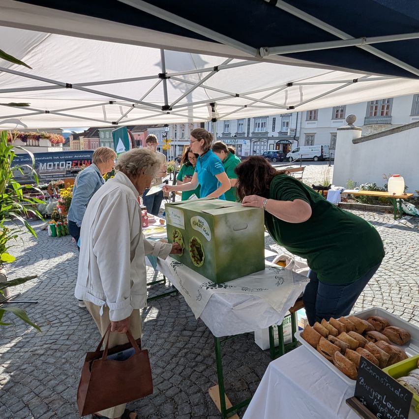 Eine Gruppe von Menschen versammelt sich unter einem Zelt bei einer Veranstaltung im Freien. Zwei Frauen untersuchen eine grüne Box, während eine andere Person eine braune Handtasche hält. Verschiedene Lebensmittel wie Brot sind auf Tischen ausgestellt. Im Hintergrund sind Gebäude und Fahrzeuge zu sehen.