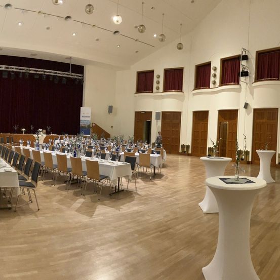 Ein großer Bankettsaal mit Holzböden und einer Bühne. Er verfügt über lange Tische mit weißen Tischdecken, leere Stühle und dekorative Elemente. Die Bühne ist mit einem Vorhang, Lautsprechern und Beleuchtungskörpern ausgestattet.
