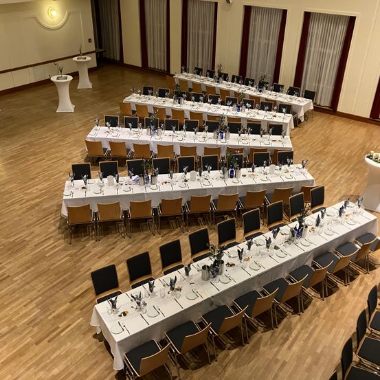 Ein Innenbankettsaal ist mit langen Tischen arrangiert, jeder mit Tellern, Besteck und Gläsern besetzt. Stühle sind ordentlich auf beiden Seiten ausgerichtet. Der Raum hat Holzböden und weiße Wände.