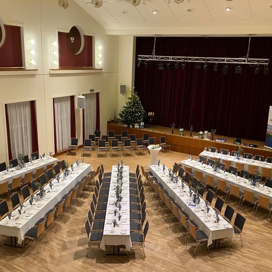 Ein großer Saal mit Holzböden, dekoriert für eine Veranstaltung. Lange Tische für ein Bankett, mit Stühlen darum herum. Eine Bühne mit einem Weihnachtsbaum und einem roten Vorhang. Scheinwerfer an der Decke.