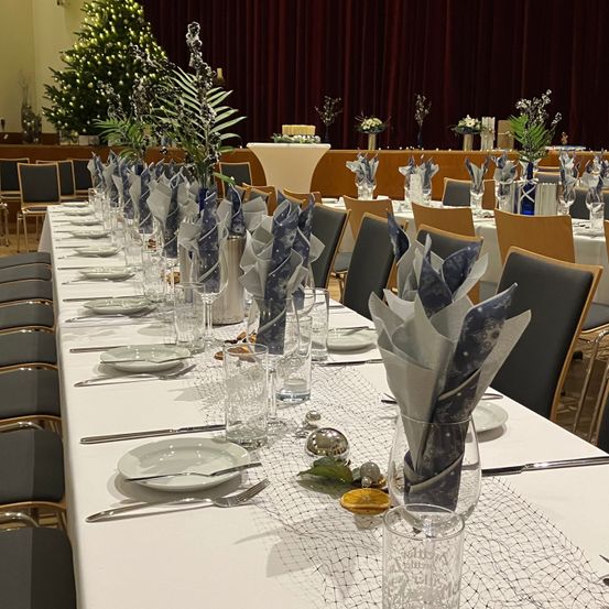 Ein langer Esstisch, der für eine Party gedeckt ist, geschmückt mit gefalteten Servietten, Gläsern und Besteck. Der Tisch ist mit einer weißen Tischdecke bedeckt und mit einem Pflanzen-Zentrenstück geschmückt. Ein Weihnachtsbaum ist im Hintergrund zu sehen.