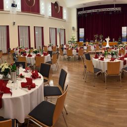 Ein Raum mit Tischen und Stühlen, die für eine Veranstaltung vorbereitet sind. Die Tische sind mit weißen Tischdecken bedeckt und die Stühle sind darum herum angeordnet. Jeder Tisch hat eine Blumendekoration, Weingläser und eine Flasche Wein. Es gibt Weihnachtsbäume und Banner im Raum.