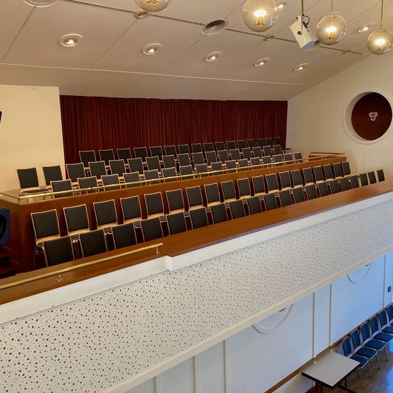 Ein leerer Hörsaal mit Reihen von Stühlen und einer Bühne mit rotem Vorhang. Der Raum hat ein modernes Design mit weißen Wänden und einer Holzbühne. In der Ecke befindet sich ein Tisch mit Monitor und Lautsprecher.
