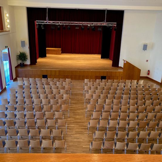 Ein leerer Saal mit einer Holzbühne, roten Vorhängen und Reihen von Stühlen. Scheinwerfer sind über der Bühne montiert.