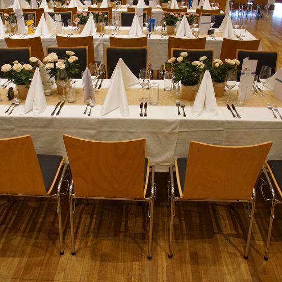 Ein gut organisierter Speisebereich mit mehreren Tischen, jeder mit weißer Tischdecke, Blumen, gefalteten Servietten und Besteck. Holzböden und Stühle vervollständigen das Ambiente.