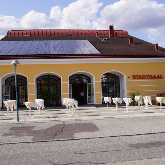 Ein gelbes Gebäude mit Solarpanelen auf dem Dach und dem Namen Stadtsaal, mit vor dem Gebäude aufgestellten Tischen und Stühlen.