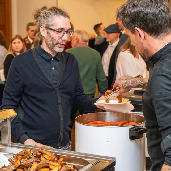 Ein Mann serviert einem anderen Mann Essen an einem Buffet. Der Mann mit Tätowierungen benutzt eine Zange, um Hot Dogs zu servieren.