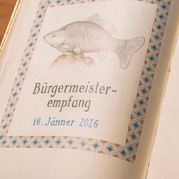Ein offenes Buch zeigt eine Zeichnung eines Fisches mit 'Burgermeister-empfang' und '16. Janner 2026' in blauem Text. Die Seite hat einen dekorativen Rand.