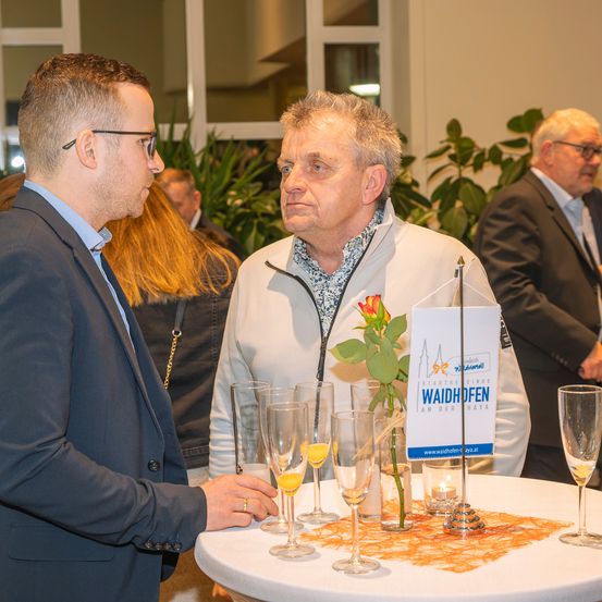 Zwei Männer unterhalten sich an einem Tisch mit Weinläsern. Ein Mann trägt eine weiße Jacke mit einer Blume und einem Namensschild. Der Tisch hat ein Namensschild, Kerzen und Weingläser. Im Hintergrund stehen andere Personen.