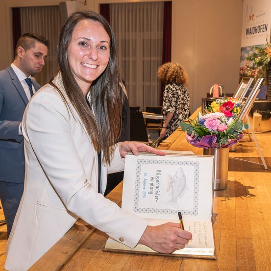 Eine Frau hält ein Buch mit dem Titel Burgermeister mit einem Stift und lächelt in einem Raum mit einem Schreibtisch, Blumen und Menschen im Hintergrund.