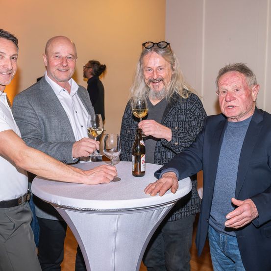 Vier Männer stehen um einen Tisch herum, lächeln und posieren für ein Foto. Zwei von ihnen halten Weingläser in den Händen, und einer hat eine Flasche Bier. Ein anderer Mann trägt eine Brille auf dem Kopf.